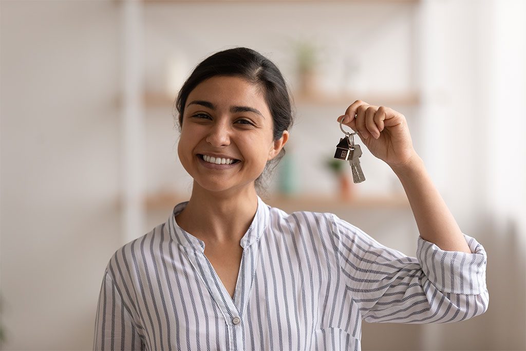 Smiling young woman holding a keyring with house-shaped keychain, suggesting she has just purchased or rented a new home.